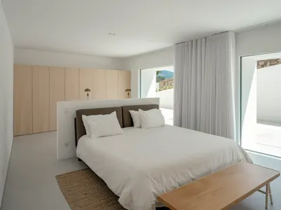 A minimalist bedroom with a large bed, white curtains, and wooden furniture.
