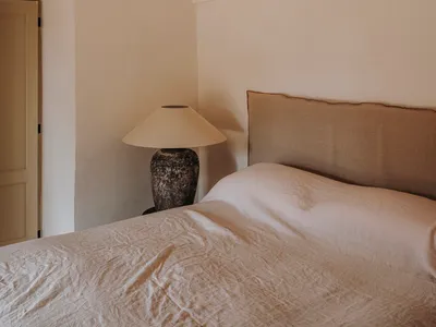 A minimalist bedroom with a beige fabric bed and a rustic lamp on a side table.