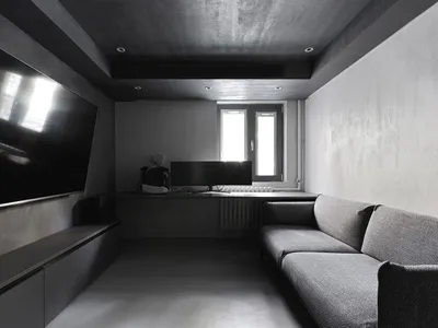 An industrial-style living room with concrete walls and floor, featuring a grey sofa and a large TV on the left wall.