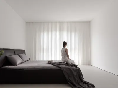 A minimalist bedroom with black furniture, grey sheets, and a woman sitting on the bed looking out through vertical blinds.
