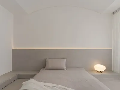Minimalist bedroom with concrete walls and floor, featuring a bed with simple bedding and a small round lamp.