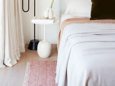 A modern minimalist bedroom features a bed with a upholstered headboard, a pink rug, and a side table with a vase of flowers.