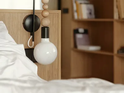 Minimalist bedroom features a white bed with wooden headboard and metal light fixture, surrounded by a bookshelf filled with