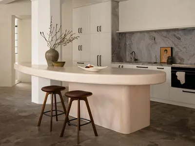 A minimalist residential kitchen featuring a concrete island with a smooth, curved countertop, two wooden stools, a vase, and