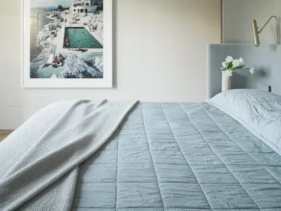 A minimalist bedroom with a light blue upholstered bed and a framed photograph of a coastal landscape.