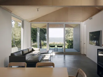 A minimalist living room with concrete flooring, a large leather sofa, and a dining area with chairs and a table.