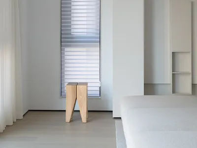 A minimalist bedroom with neutral-colored walls, floor, and bedding, featuring a modern light-colored wooden stool and a larg