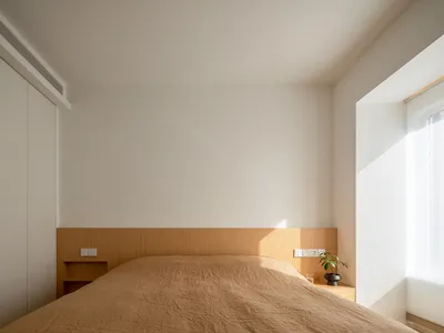 Minimalist bedroom featuring a wooden bed with a plain wall and a window with blinds, highlighting a serene and modern aesthe