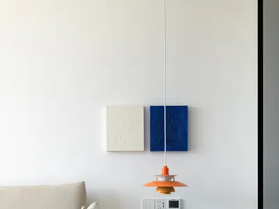 A minimalist bedroom with a bed, pillows, and a hanging lamp against a white wall with framed art.