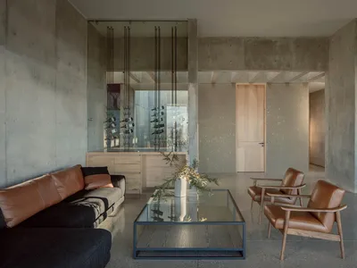 Modern industrial living room with concrete walls, a glass coffee table, leather armchairs, and a wine rack.
