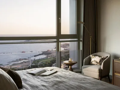 Modern minimalist bedroom with a bed, chair, and table by a large window overlooking the ocean.