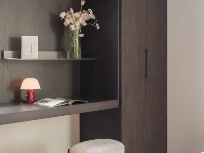 A minimalist bedroom featuring wooden furniture, a bed, a desk with a lamp and flowers, and a shelf.