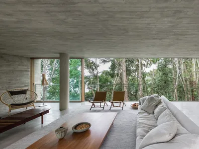 Industrial-style living room with concrete floor, minimalist chairs, and large windows overlooking a forest.