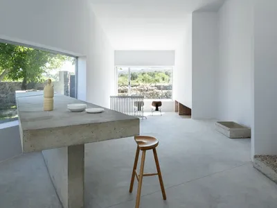 A modern minimalist kitchen featuring a concrete table and stool, surrounded by white walls and large windows with a view of