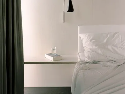 A minimalist bedroom with a white bed and grey curtains, featuring a white nightstand, a fabric lampshade, and a glass water