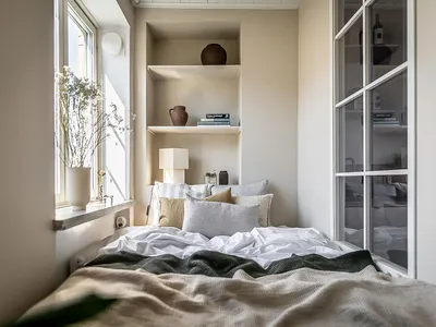 A cozy, minimalist bedroom with neutral-colored bedding, shelves filled with books and vases, and a window letting in natural