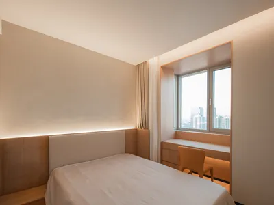 Minimalist bedroom with a white bed and wooden accents, featuring a window with a city view.