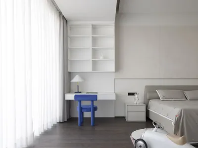 Modern, minimalist bedroom featuring a white bed with neutral bedding, a blue chair, and white shelves with a lamp on a desk.