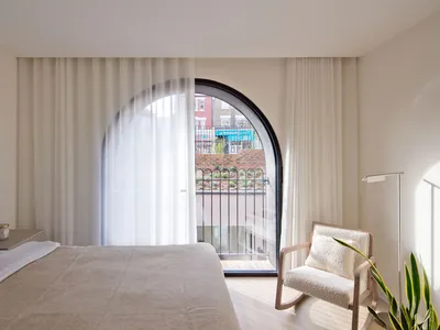 A minimalist bedroom with a large arched window, a bed with light-colored bedding, and a rocking chair.