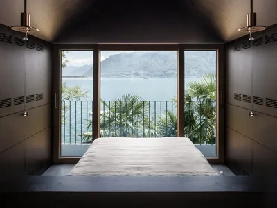 Minimalist bedroom featuring a bed with a white sheet against a window with a scenic view of mountains and a lake.