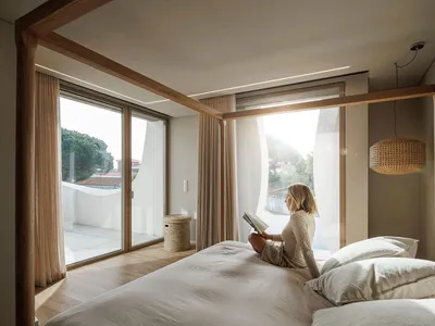 A modern minimalist bedroom with natural wooden elements, featuring a woman reading a book seated on the bed, overlooking a b