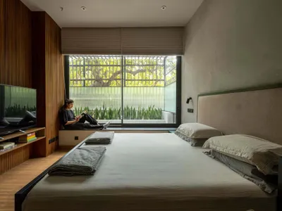 A modern minimalist bedroom featuring a large bed, wooden walls, and fabric bedding, with a person sitting on a window sill r
