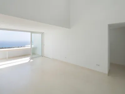An empty minimalist living room with high ceilings, large glass windows overlooking the ocean, and white walls and floor.