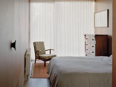 A minimalist bedroom with a white flat door, sheer white curtains, and a large window, featuring a bed with a white bedspread