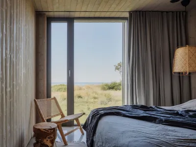 A serene minimalist bedroom with a large window overlooking a natural landscape, featuring a wooden bed, chair, and stool, an