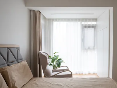 A minimalist bedroom featuring a beige upholstered bed, a modern beige armchair, light wood flooring, and sheer curtains on a