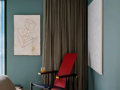 A modern bedroom featuring fabric curtains, a vibrant red and blue reclining chair, and geometric abstract art on the walls.