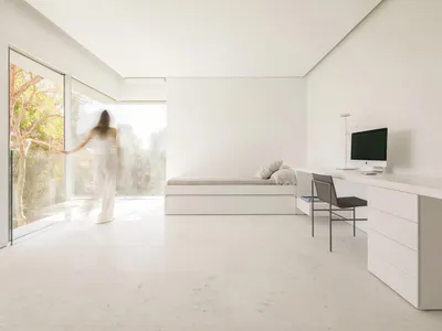 一間極簡風格的臥室，配有內建收納的流線型現代床、簡潔的白色書桌，以及大面玻璃窗。