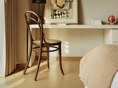 Minimalist bedroom with a wooden chair, a desk with a framed abstract painting, and a bed covered in beige textiles.