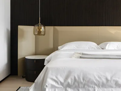 A modern minimalist bedroom with a beige headboard, white bedding, and a dark wood accent wall.