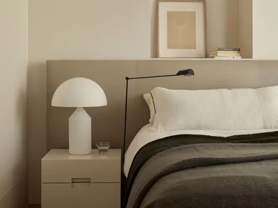 A minimalist bedroom with a white bed, a nightstand with a white lamp, and a floating shelf above the bed.