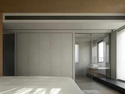 Modern minimalist bedroom with neutral-toned walls, wardrobe, and bed, featuring a bathroom with glass partition.