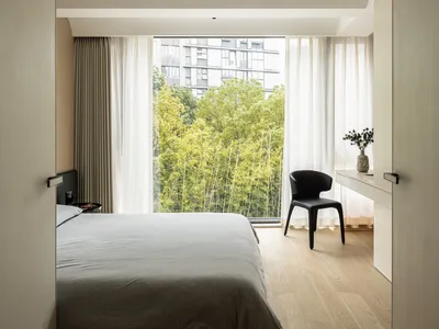 Bedroom with minimalist design, featuring a bed with wood headboard and a chair, natural light from large windows, and wooden