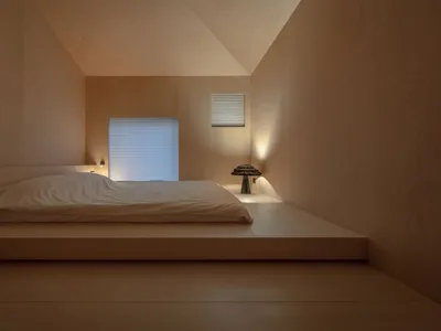 A minimalist bedroom with a wood floor, bed, and window with blinds.