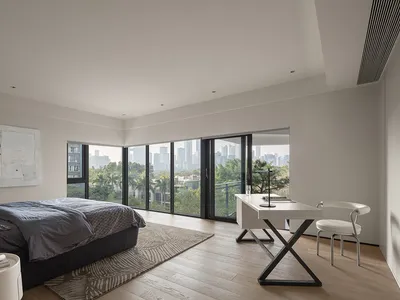 A modern minimalist bedroom featuring a large bed with dark bedding, a white desk, and floor-to-ceiling windows overlooking a