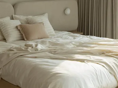 A minimalist bedroom with a bed dressed in neutral colors, including white sheets, a beige throw pillow, and a beige headboar