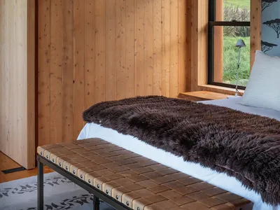 A minimalist bedroom with wooden walls, a fur blanket, and a window overlooking a green landscape.