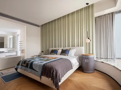 A modern minimalist bedroom with a light wooden floor, white walls, and a large bed with geometric patterned bedding.