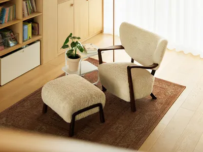 A cozy living room with a comfortable armchair and matching ottoman, wooden flooring, and a side table with a plant and books