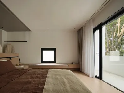 A minimalist bedroom featuring a bed with brown and beige bedding, a wooden headboard, and a window with white curtains.