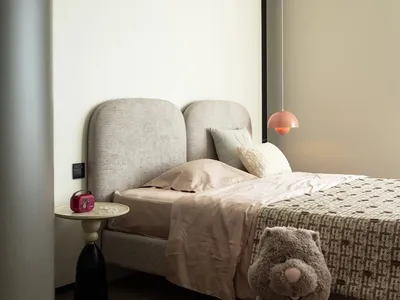 A modern minimalist bedroom featuring a gray upholstered headboard, pink pendant lamp, and a large teddy bear on the floor.