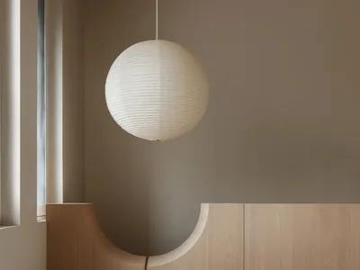A minimalist bedroom featuring a wooden bed with a pendant light above it, set against a neutral wall and a window with blind