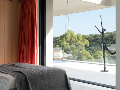 A minimalist bedroom with concrete flooring, a grey blanket, and a large window overlooking green trees.