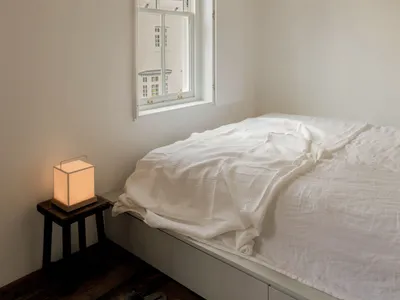 A minimalist bedroom with a white bed, wooden floor, and natural light coming through the window.