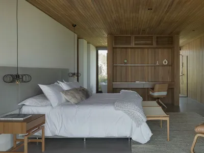 A modern monochromatic bedroom featuring white bedding and a wooden floor with a large bed, wooden shelves, and a wooden chai