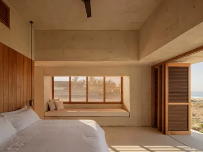 A minimalist bedroom with concrete walls, a bed with white bedding, and cushions on a window seat.